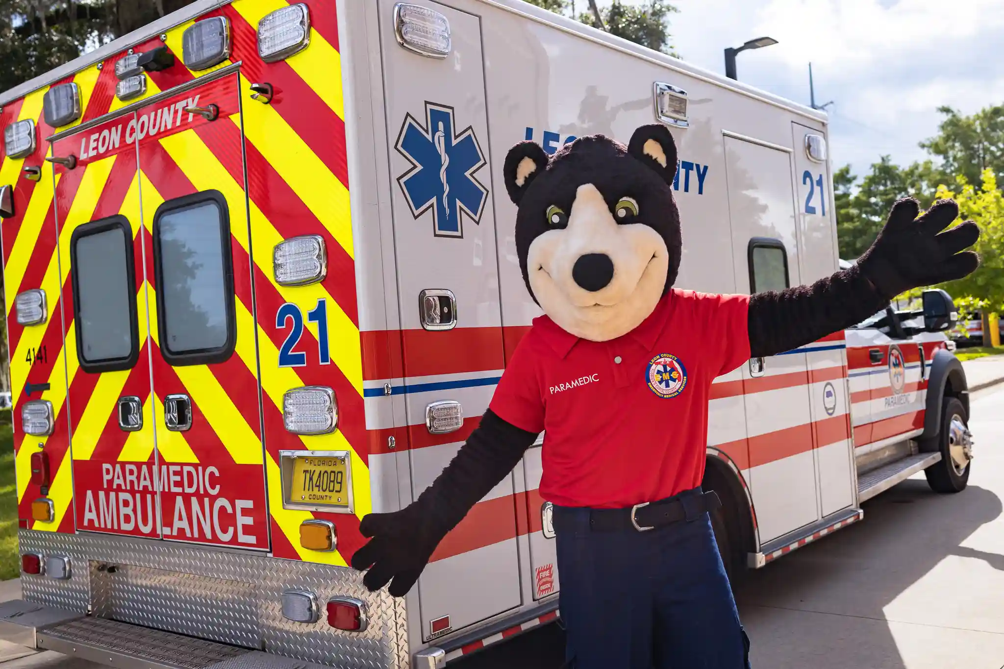 Black bear mascot with a red Leon County EMS shirt posing in front of an EMS ambulance. 